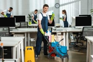 cheerful cleaner moving cart with cleaning supplies near multicultural colleagues