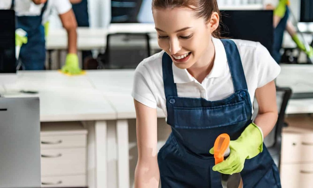 cheerful cleaner in overalls cleaning office desk with rag
