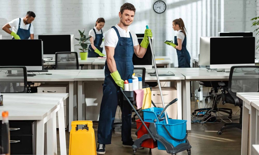 cheerful cleaner moving cart with cleaning supplies near multicultural colleagues