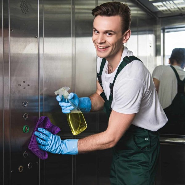 young cleaning company worker cleaning elevator and smiling at camera