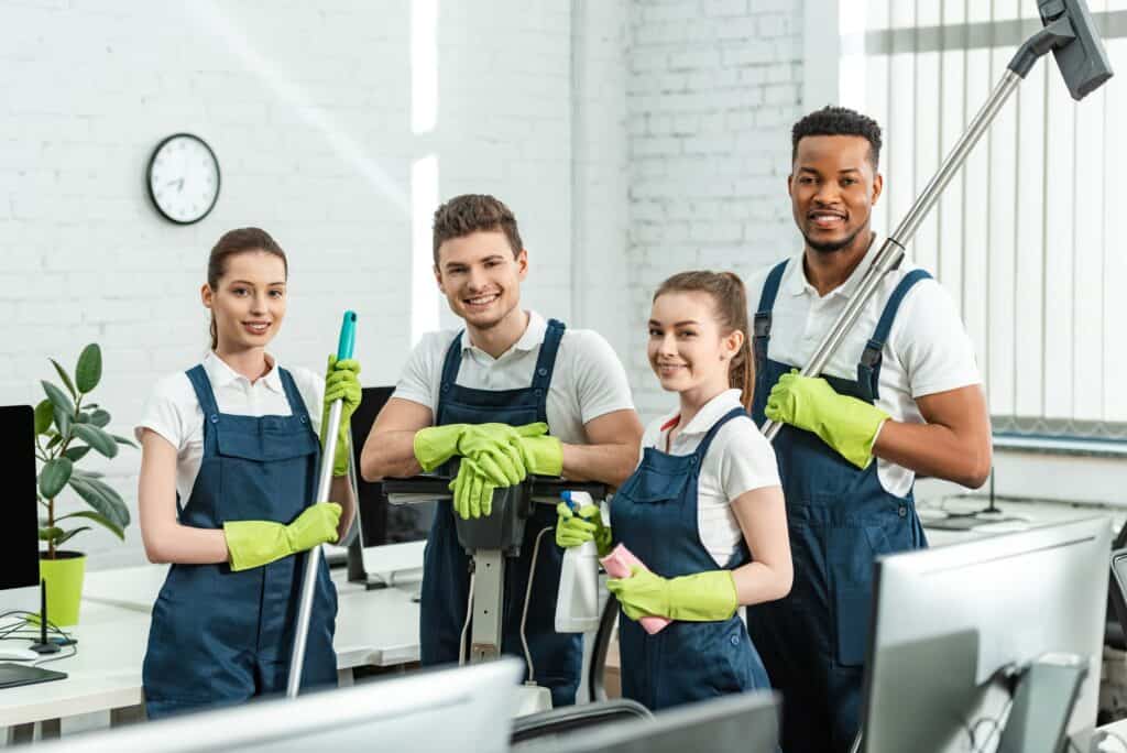 happy multicultural cleaners looking at camera while standing with cleaning supplies in office