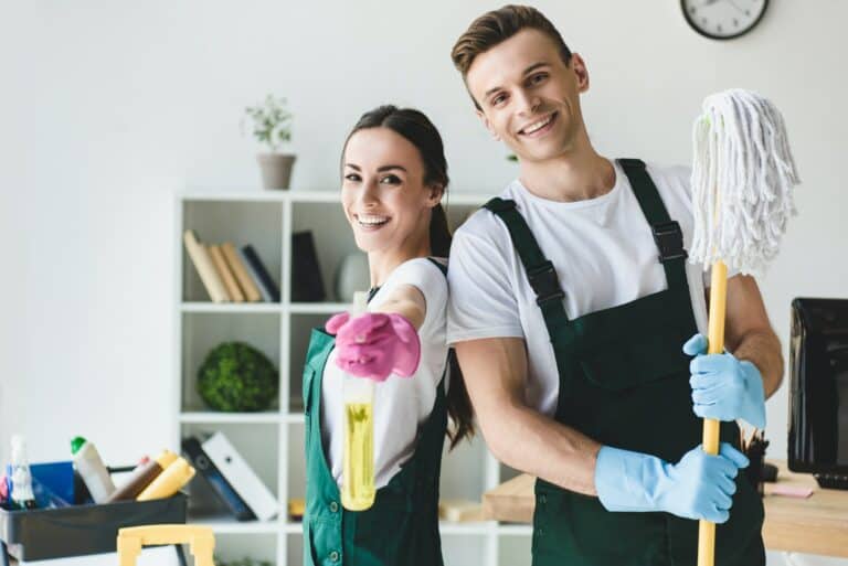 happy young cleaners with mop and spray bottle smiling at camera while cleaning office