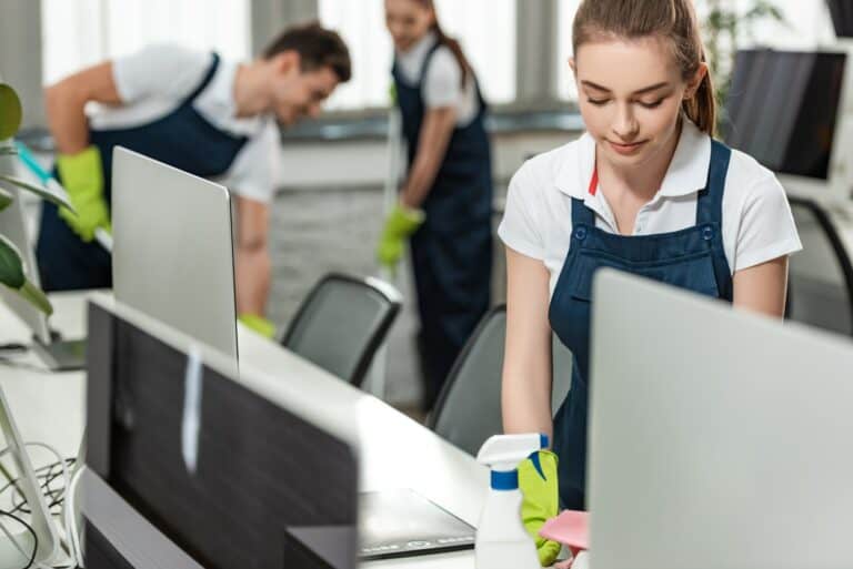 selective focus of pretty cleaner wiping desk while colleagues cleaning floor in office