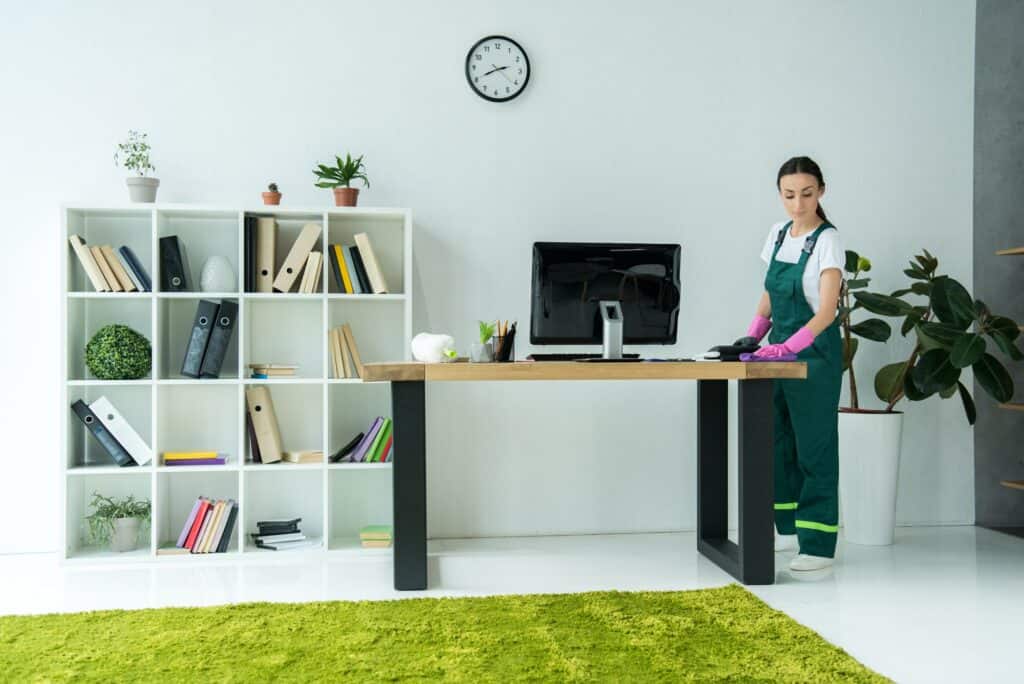 young cleaning company worker in rubber gloves and uniform cleaning desk in modern office